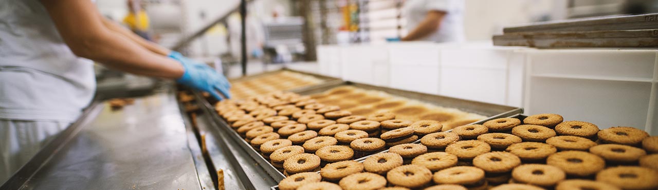 Persoon met persoonlijke beschermingsmiddelen aan de lopende band in een koekjesfabriek.